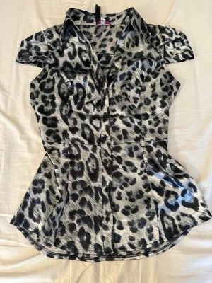 Leopardmönstrad kortärmad blus - Snygg blus med leopardmönster i svart, grått och vitt. Modellen har korta ärmar med veckade detaljer, figurnära passform och klassisk krage. Materialet är glansigt och ger en lyxig känsla. Perfekt för dig som vill sticka ut med ett djurmönster.