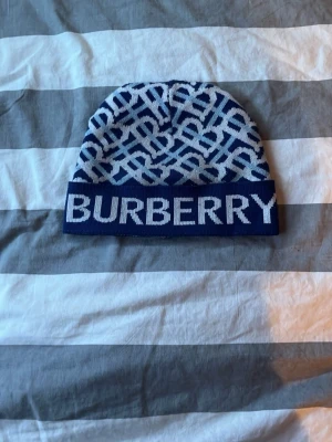 Blåvit Burberry mössa med logga - Säljer en snygg blåvit mössa från Burberry med stort logotyp-tryck framtill och ett grafiskt mönster över hela mössan. Tillverkad i mjukt stickat material som håller dig varm och stilsäker under kalla dagar.
