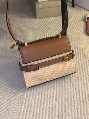 Brun och beige handväska i läder och rotting - Strukturerad hand-/axelväska i naturfärgat flätat material med bruna detaljer. Kompakt men stadig modell med lock och dekorativa spännen framtill. Avtagbar och justerbar axelrem. Perfekt storlek för vardagsbruk när du vill bära lätt men ordnat.🤍  Structured handbag/shoulder bag in natural woven material with brown details. Compact yet sturdy shape with a flap closure and decorative front buckles. Detachable and adjustable shoulder strap. Ideal size for everyday use when you want something light 