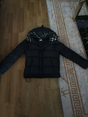 Svart pufferjacka Burberry med avtagbara ärmar - Snygg svart pufferjacka från Burberry med avtagbara ärmar så den kan användas som väst. Jackan har huva med klassiskt rutigt foder, dragkedja och knappar framtill samt två fickor med dragkedja. Perfekt för kalla vinterdagar.