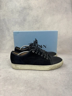 Svarta sneakers från Lanvin - Stilrena svarta sneakers från Lanvin med ovandel i mocka och tåparti i slätt läder. Skorna har vita, slitna sulor och svarta skosnören. Klassisk låg modell med metallöljetter och diskret logga på sidan.