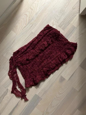 Minikjol - Supersöt vinröd minikjol med volangdetaljer. Kjolen har en knytning i sidan och är gjord i ett lätt, skirt material som ger en romantisk vibe. Perfekt att styla med en enkel topp för en trendig look. Spets. Oanvänd.