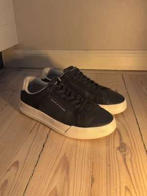 Svarta Tommy Hilfiger sneakers - Snygga svarta sneakers från Tommy Hilfiger med vit sula och diskret logga på sidan. Skorna har klassisk låg profil, svarta skosnören och ovandel i läderliknande material. Perfekta för en clean och stilren look.