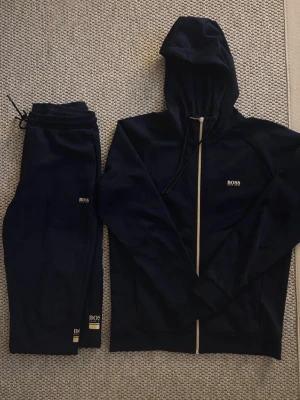 Mörkblå BOSS set - Snyggt mörkblått loungewear set från BOSS med matchande hoodie och byxor. Hoodien har dragkedja, justerbar huva och BOSS-logga på bröstet. Byxorna har resår i midjan och logga på benet. Perfekt för chill dagar hemma.