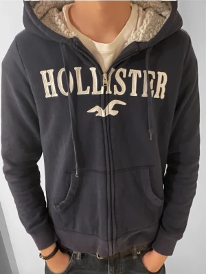 Mörkblå hoodie från Hollister - Mysig mörkblå hoodie från Hollister med dragkedja och vit broderad logga framtill. Fodrad med mjuk teddy på insidan av huvan och har snörning samt två fickor framtill. Perfekt för en avslappnad stil. Det står storlek M men passar bättre som storlek S. Modellen är 180 och väger 65.