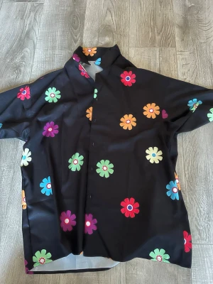 Svart blommig skjorta från Manfinity - Säljer en svart skjorta från Manfinity med stora färgglada blommor i rött, blått, grönt, orange, lila och vitt. Skjortan har klassisk krage och knäppning framtill. Perfekt för dig som gillar färg och vill sticka ut lite extra.