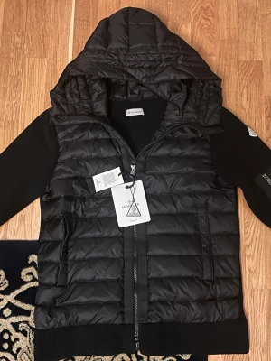 Moncler cardiganjacka i storlek M - Helt Ny - Denna Moncler cardiganjacka i storlek M kombinerar funktion och stil. Den har en quiltad design med en huva för extra värme och komfort. Perfekt för kalla dagar, och med sin svarta färg passar den både till vardags och mer avslappnade tillfällen. Jackan har också en praktisk armficka och en trendig siluett som gör den till ett utmärkt val för både stilmedvetna och praktiska användare.