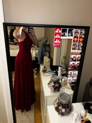 Vinröd balklänning med spetsdetaljer - Säljer en vinröd balklänning med spets upptill. Klänningen har en elegant, flytande kjol och markerad midja. Perfekt för bal eller festliga tillfällen. Materialet känns lätt och faller snyggt.