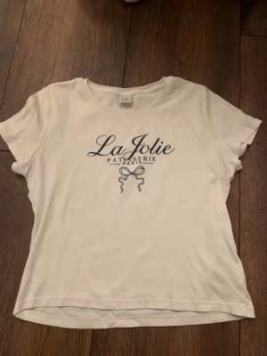 T-shirt med tryck  - Jag säljer en kortärmad T-shirt från Lindex. Tröjan har ett tryck med text och en rosett. Jag svarar gärna på era frågor.🫶🏻🫶🏻