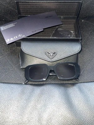 Svarta solglasögon från Prada - Snygga svarta solglasögon från Prada med klassisk design. Kommer med originalfodral och äkthetskort. Perfekta för dig som vill ha en stilren accessoar med lyxig känsla.