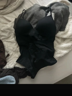 Svart shaping-body med tunna band - Snygg svart shaping-body med tunna justerbara axelband och vadderade kupor. Bodyn har en slät, figurnära passform och är tillverkad i ett stretchigt material som formar kroppen. Perfekt under tighta plagg för en smickrande siluett.