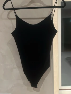 Svart basic body med smala axelband - Enkel och stilren svart body med smala axelband och rundad halsringning. Perfekt att matcha med jeans eller kjol för en clean look. Materialet känns stretchigt och följsamt. Inga synliga detaljer eller mönster. Lite smutsig bak men försvinner i tvätt