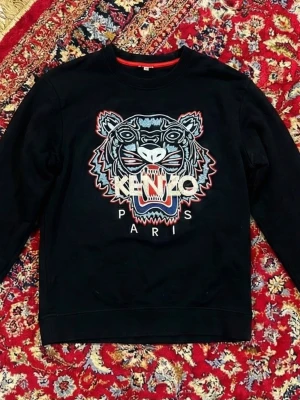 Svart Kenzo sweatshirt med tigertryck - Cool svart sweatshirt från Kenzo med ikoniskt broderat tigerhuvud och texten 'KENZO PARIS' på bröstet. Tröjan har rund halsringning och är tillverkad i 100% bomull. Perfekt för dig som gillar streetwear och vill sticka ut.