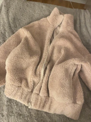 Beige teddyjacka med dragkedja - Supermjuk och fluffig teddyjacka i beige färg med hög krage och dragkedja framtill. Jackan har två stora fickor och en avslappnad passform som ger en cozy vibe. Perfekt för kyliga dagar när du vill vara både varm och stylish.