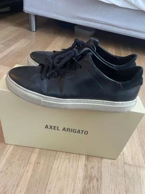 Svarta Axel Arigato sneakers i läder - Stilrena svarta sneakers från Axel Arigato i mjukt läder med svart snörning och vit sula. Klassisk låg modell med minimalistisk design och diskret logga på sidan. Perfekta för dig som gillar clean och modern stil. Man får med lådan