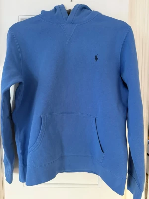 Blå hoodie från Ralph Lauren - Snygg blå hoodie från Ralph Lauren med klassisk huva och känguruficka framtill. Liten mörkblå logga broderad på bröstet. Tillverkad i mjukt bomullsmaterial som är skönt att ha på sig. Perfekt för en avslappnad och stilren look.