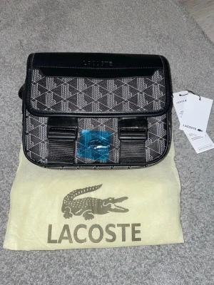 Svart axelväska från Lacoste - Snygg svart axelväska från Lacoste med geometriskt mönster i vitt och svart. Väskan har justerbar axelrem, lock med spännen och detaljer i konstläder. Perfekt för dig som vill ha en stilren och modern accessoar med tydlig logga. Pris kan självklart diskuteras!