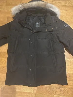 Svart dunjacka från Canada Goose - Snygg svart dunjacka från Canada Goose med avtagbar pälskant på huvan och klassisk logga på ärmen. Jackan har dragkedja och knappar framtill samt flera praktiska fickor. Perfekt för kalla dagar och riktigt varm.