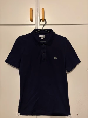 Marinblå pikétröja från Lacoste - Klassisk marinblå pikétröja från Lacoste med slim fit-passform. Tröjan har korta ärmar, krage och knappar framtill samt den ikoniska gröna krokodilloggan på bröstet. Perfekt till jeans eller shorts för en clean och stilren look.