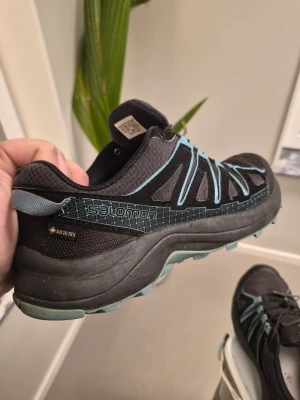 Salomon Löparskor, Svart, EUR 38 2/3 - Säljer ett par svarta Salomon kängor med blå detaljer och snabbsnörning. Skorna är tillverkade i slitstarkt syntetmaterial och har Gore-Tex-membran som håller fötterna torra. Perfekta för utomhusaktiviteter och äventyr.