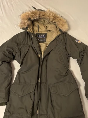 Olivgrön parkas med fuskpäls från Svea - Olivgrön parkas från Svea med stor huva och fuskpäls. Jackan är figurnära och har inga defekter förutom hängbandet som lossnat! Säljer den då den inte kommer åt användning 🥰