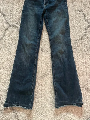 Mörkblå bootcut jeans med råa kanter - Säljer ett par mörkblå bootcut jeans med fransade, råa kanter i midja och benslut. Jeansen har klassisk femficksdesign och är gjorda i denim med lätt tvättade detaljer. Perfekta för en avslappnad och trendig look.