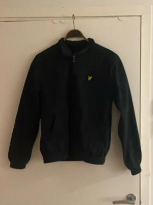 Mörkblå bomberjacka från Lyle & Scott - Klassisk mörkblå bomberjacka från Lyle & Scott med gul logga på bröstet. Jackan har dragkedja framtill, ribbade muddar och krage. Insidan har ett rutigt foder i färgglada toner. Perfekt för dig som vill ha en stilren och tidlös look.