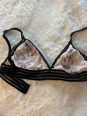 Bralette med broderade fåglar - Oanvänd bralette i svart mesh med vita kupor dekorerade med färgglada broderade fåglar och blommor. Justerbara axelband och bred resår under bysten.