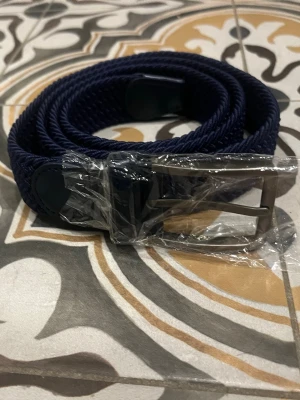 Mörkblå flätat bälte med metallspänne - Snyggt mörkblått bälte med flätad design och ett stilrent metallspänne. Perfekt accessoar för att lyfta din outfit. Bältet har detaljer i syntetmaterial vid spännet och ändarna. Helt nytt