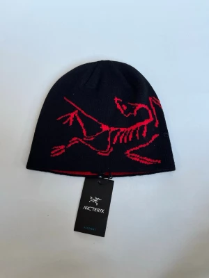 Svart mössa från Arc'teryx med röd logga - Snygg svart mössa från Arc'teryx med röd dinosaurielogga. Tillverkad i en lyxig blandning av 49% kashmir, 49% ull och 2% nylon. Mössan har en tight passform och är perfekt för kalla dagar.