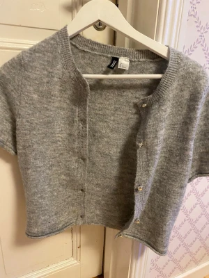Grå kortärmad kofta från H&M - Kortärmad grå kofta från H&M i mjukt stickat material. Modellen är croppad med rund halsringning och knappar framtill. Perfekt att styla över ett linne eller t-shirt för en trendig lager-på-lager-look.