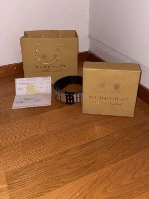 Burberry rutig bälte med svart spänne - Säljer ett klassiskt Burberry bälte med det ikoniska rutiga mönstret i beige, svart och rött. Bältet har ett svart metallspänne med Burberrys logga. Perfekt accessoar för att lyfta din outfit.