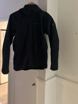 Svart vindjacka från Peak Performance - Snygg svart vindjacka från Peak Performance med huva och dragkedja framtill. Jackan har två sidofickor med dragkedja och en minimalistisk logga på bröstet. Perfekt för blåsiga dagar och enkel att matcha med allt.