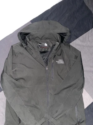 Svart vindjacka från The North Face - Snygg svart vindjacka från The North Face med huva och dragkedja framtill. Jackan har två sidofickor med dragkedja och en bröstficka. Loggan är tryckt på både bröstet och ryggen. Perfekt för blåsiga dagar och enkel att matcha.