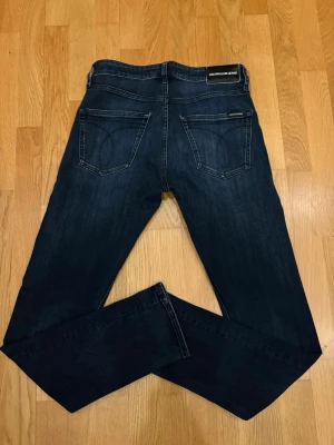 Mörkblå slim jeans Calvin Klein - Snygga mörkblå slim fit jeans från Calvin Klein Jeans med klassisk femficksmodell och subtila slitningar framtill. Jeansen har raka ben och logotypdetaljer på bakfickan och linningen. Perfekta för en clean och modern look.