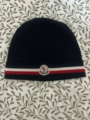 Mörkblå Moncler mössa med ränder - Snygg mörkblå mössa från Moncler med ribbstickad kant och klassiska ränder i vitt och rött. Framtill sitter Monclers ikoniska logotyp. Perfekt accessoar för att hålla värmen och addera stilpoäng till din outfit.