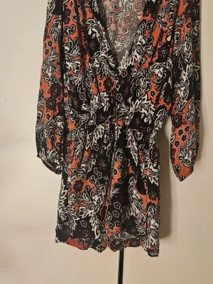 Blommig byxdress i orange och svart - Snygg byxdress med blommigt mönster i svart, vitt och orange. Byxdressen har markerad midja med knytband och djup v-ringning. Lätt och luftig i materialet, perfekt för dig som gillar boho-vibbar och färgstarka prints.