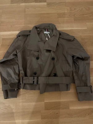 Kort beige trenchcoat Tommy Hilfiger - Snygg kort beige trenchcoat från Tommy Hilfiger med dubbelknäppning, axelklaffar och bälte i midjan. Jackan har klassisk krage, justerbara ärmslut och är tillverkad i ett glansigt, slitstarkt material. Perfekt för en trendig och stilren look. Använd 1 gång.
