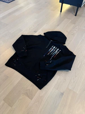 Svart hoodie från Amiri med färgstänk - Säljer en svart hoodie från Amiri med stor huva och coola färgstänk i vitt, rött och blått. På ryggen står det AMIRI i stora bokstäver med drip-effekt. Hoodien har en avslappnad passform och känns riktigt street. Perfekt för dig som vill sticka ut.