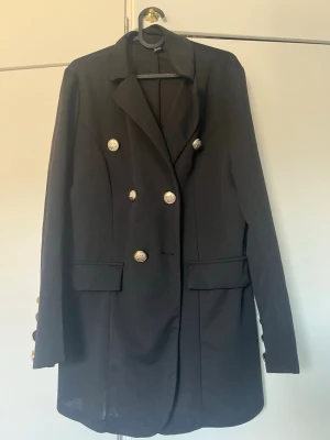 Svart kavaj med guldknappar - Stilren svart kavaj med dubbelknäppning och dekorativa guldknappar framtill och vid ärmsluten. Kavajen har två fickor med lock och en klassisk krage. Perfekt för en trendig och elegant look.