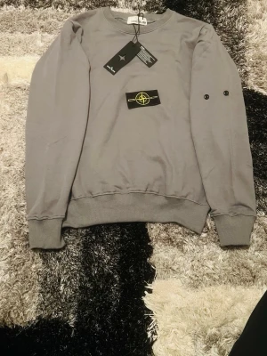 Grå sweatshirt från Stone Island - Säljer en stilren grå sweatshirt från Stone Island med klassisk rund hals och ribbade muddar. Tröjan har det ikoniska Stone Island-märket som kan fästas på ärmen med knappar. Perfekt för dig som gillar streetwear och enkel design.