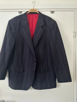 Rutig Hugo Boss kavaj / blazer, XL - Klassisk marinblå / navy / blå kavaj / blazer från Hugo Boss i storlek XL (se exakta mått längre ned). Stiligt rutmönster i rött och beige, och glansigt lent foder i klarrött med Hugo Boss-loggan. Två fodrade fickor på utsidan och flertalet smarta fack och fickor på insidan. Mycket gott skick!  MÅTT (tvärsöver plagget): Bystmått ca 58cm, midjemått ca 55cm, axelbredd ca 49cm, längd ca 74cm, ärmlängd ca 61cm.   Postas mot porto eller hämtas i Stockholm.