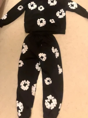 Svart loungewear-set med vita blommor - Säljer ett svart loungewear-set med matchande tröja och byxor, båda dekorerade med stora vita blommönster. Tröjan har långa ärmar och byxorna har resår i midjan och vid bensluten. Perfekt för chill dagar hemma.