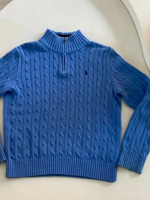Blå kabelstickad tröja Polo Ralph Lauren äkta! - Snygg blå kabelstickad tröja från Polo Ralph Lauren med halv dragkedja och ribbad krage med svarta ränder. Tröjan har lång ärm, ribbade muddar och den klassiska broderade loggan på bröstet. Perfekt för dig som gillar stilren och sportig look.