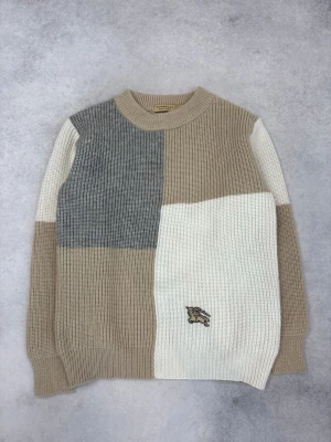 Blockmönstrad stickad tröja Burberry - Snygg stickad tröja från Burberry i blockmönster med beige, grå och vit färg. Tröjan har rund halsringning och en broderad logga framtill. Grovstickad i mjukt material, perfekt för lager-på-lager-stil.