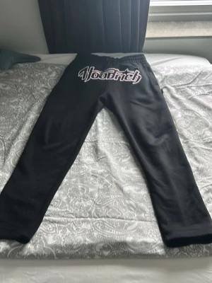Svarta Hoodrich mjukisbyxor - Säljer ett par svarta mjukisbyxor från Hoodrich med stor logga i vitt och rött framtill. Byxorna har resår i midjan med dragsko och raka ben. Perfekta för chill dagar och streetwear-stil.
