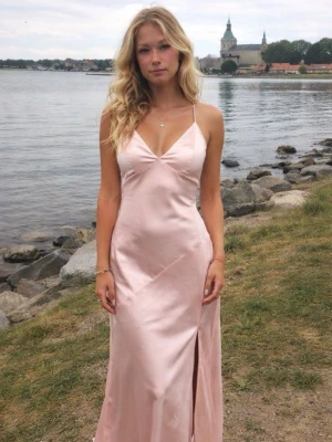 Ljusrosa lång satinklänning - Säljer en ljusrosa lång klänning i satin med smala axelband och djup v-ringning. Klänningen har öppen rygg med korsade band och en slits framtill. Perfekt för bal eller sommarfest. Materialet är glansigt och följsamt. Storlek 36, men skulle säga att de passar 34 med!