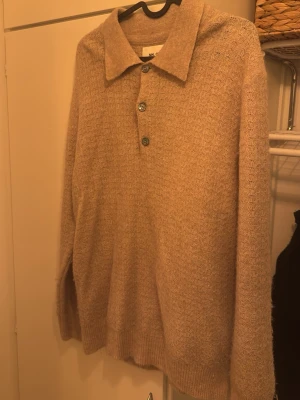 Beige stickad tröja från NN07 - Stickad tröja från NN07 i beige med diskret mönster, klassisk krage och tre knappar framtill. Tröjan har lång ärm och ribbade muddar. Perfekt för dig som gillar stilrena och bekväma plagg i mjukt material. Svår att ta bild på men väldigt fint stickad. 