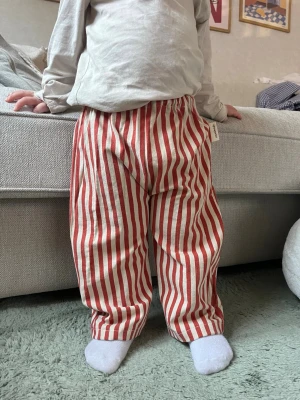 Randiga byxor från Handmade - Säljer ett par lösa byxor med röda och vita vertikala ränder från Handmade. Byxorna har resår i midjan och är tillverkade i ett mjukt, vävt bomullstyg. Perfekta för en avslappnad och trendig stil.