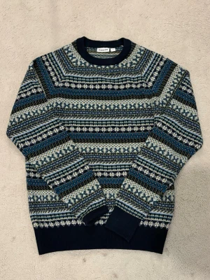 Mönstrad stickad tröja från Kappahl - Stickad tröja från Kappahl i storlek S med klassiskt mönster i blått, vitt, grönt och svart. Tröjan har rund halsringning, ribbade muddar och en mysig, retroinspirerad look. Perfekt för kalla dagar och snygg till jeans.